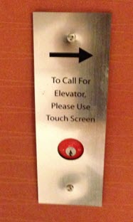 Metal sign with text that says to call for elevator please use touch screen but arrow is pointed to the right.
